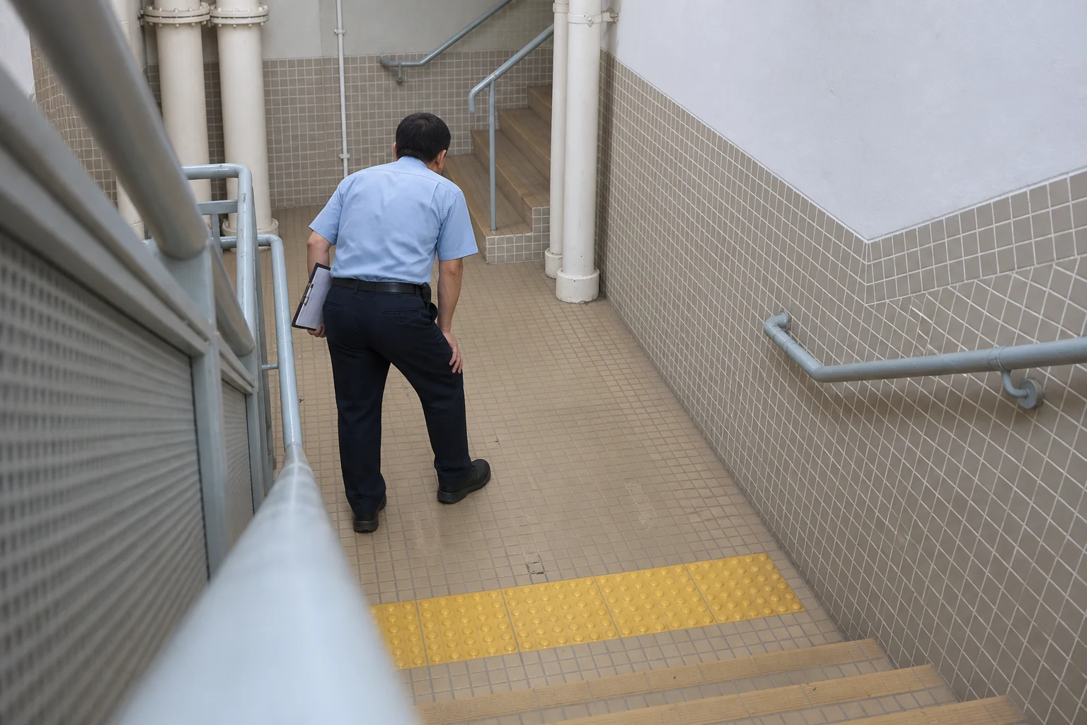 A security guard seen from behind, resting a hand on his bent knee in a stairwell; no face visible. AI illustration based on Stairio field imagery.